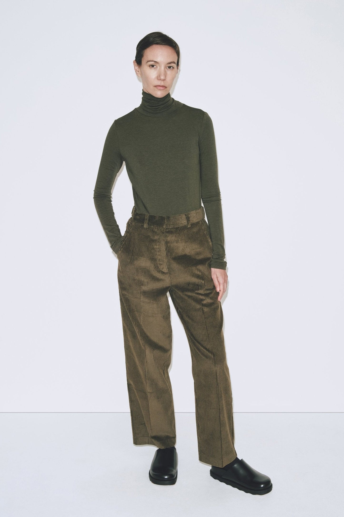 A person wearing a green roll neck jersey top with long sleeves and slim fit, paired with brown trousers.