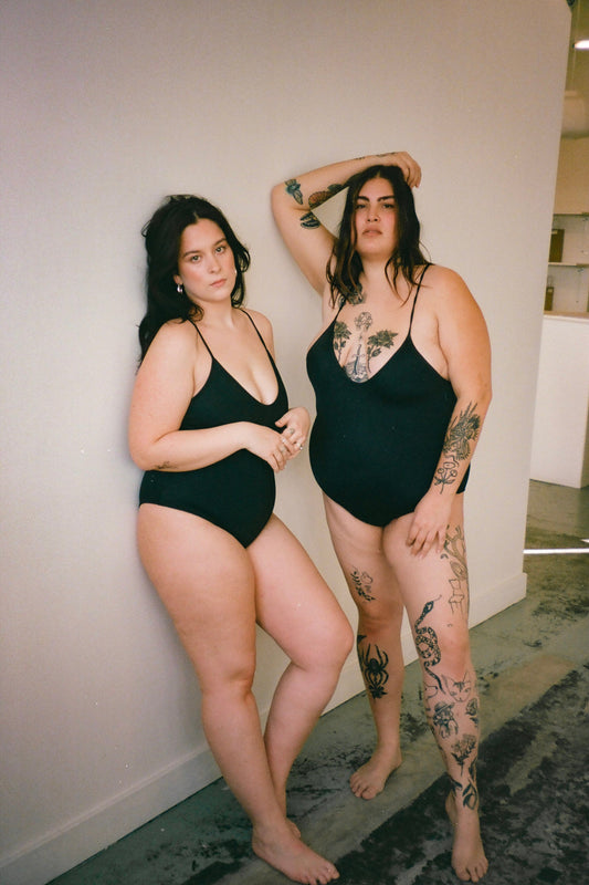 Two models wearing the Hattie One-Piece swimsuit in black. The swimsuit has a scoop neckline, cheeky coverage, and low back with adjustable crisscross spaghetti straps.