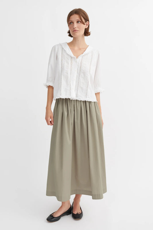 A woman wearing a white top and a green, wide organic cotton skirt with an elastic waistband, standing against a plain background.