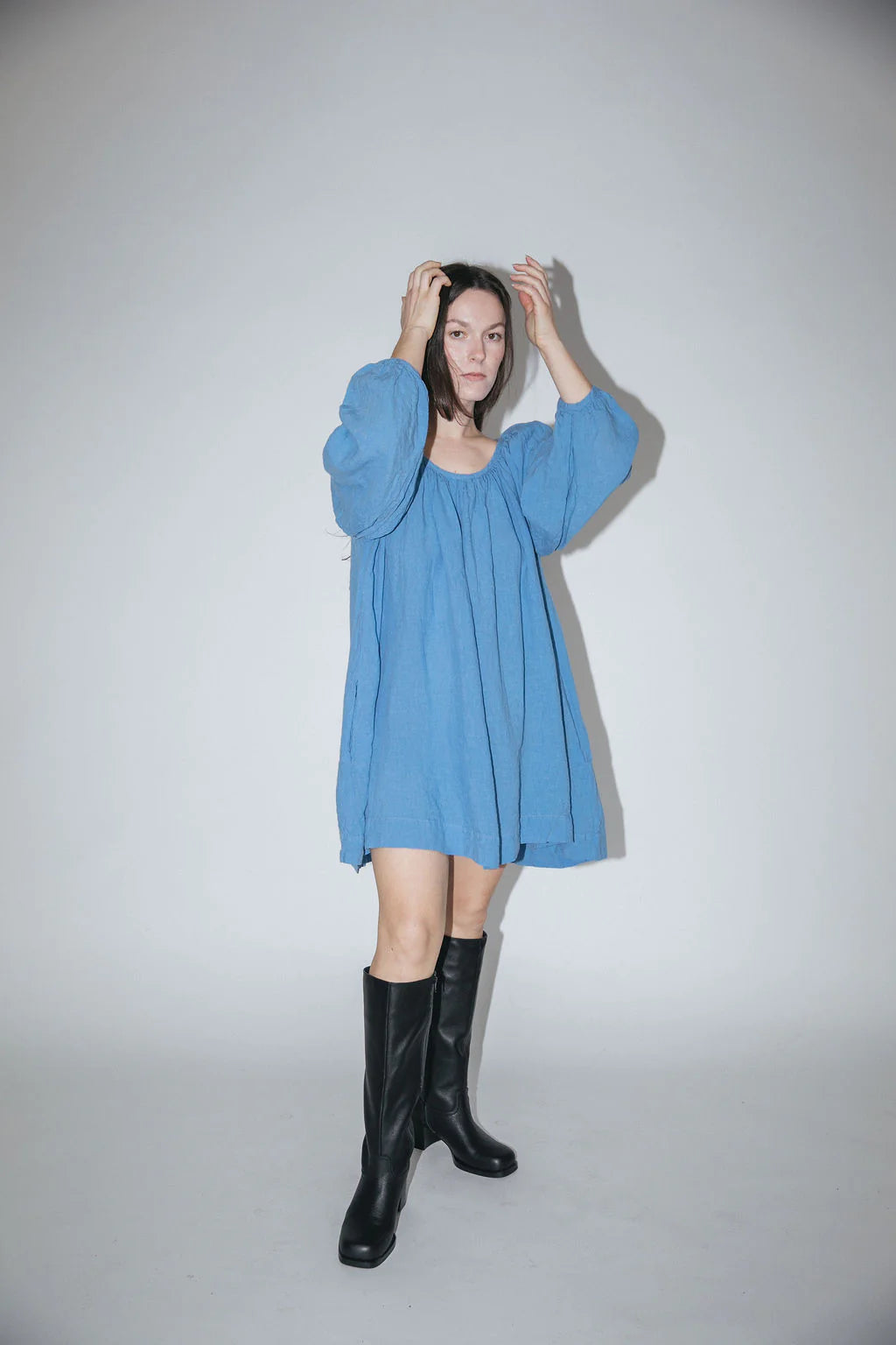 A person standing against a white background, wearing a blue mini dress with puff sleeves and a gathered neckline, paired with black boots.