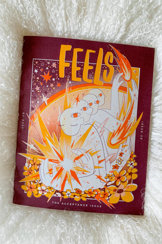 A colorful magazine cover of 'Feels' with illustrated text and graphics on a purple background, placed on a fluffy white surface.