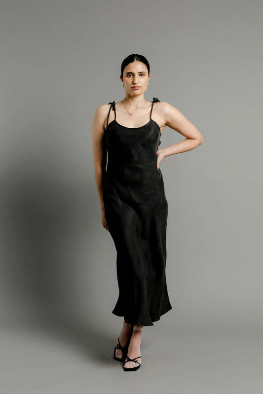 A woman standing in a studio setting, wearing a black sleeveless slip dress with adjustable straps and a V-neckline.