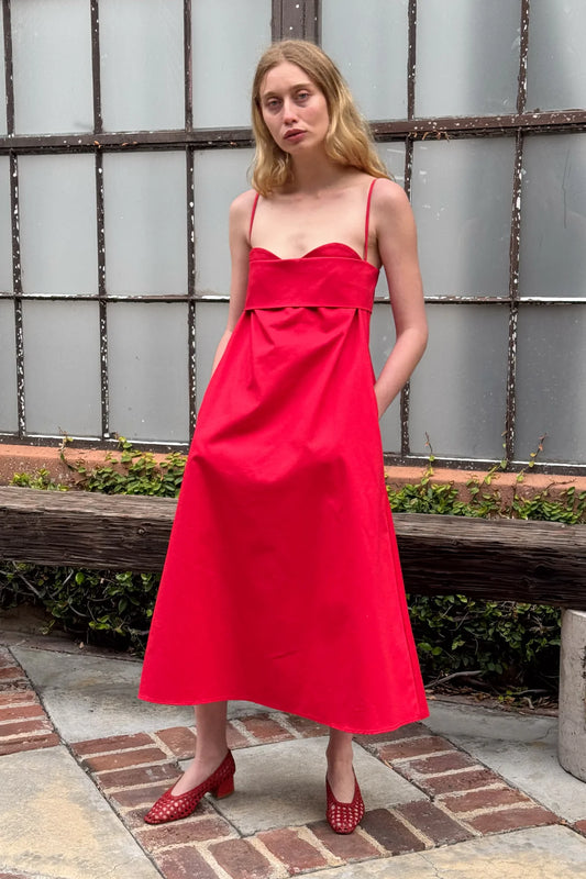 A woman standing outdoors wearing a red, V-neck, short-sleeved dress with a sweetheart neckline, made from organic cotton twill, posing against a backdrop of a building with glass panels.