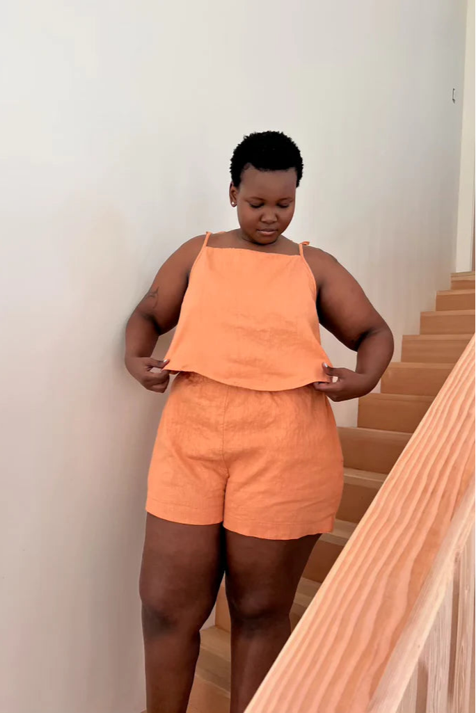 A person standing on stairs wearing an orange linen tank top and matching shorts. The tank top has adjustable tie closures at the shoulder.