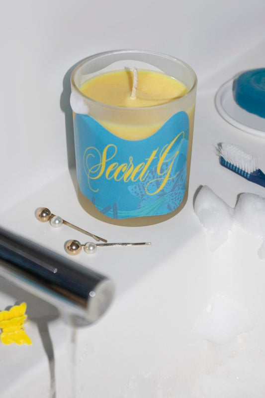 A candle labeled 'Secret G' with a blue and yellow design, placed on a bathroom counter next to a toothbrush and other bathroom accessories.