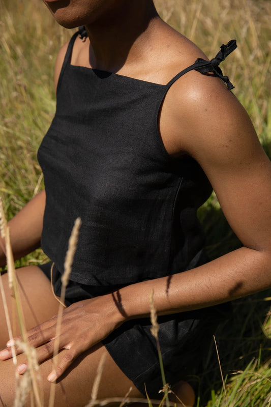 A person sitting in a field wearing a black sleeveless linen tank top with adjustable tie closures at the shoulder.
