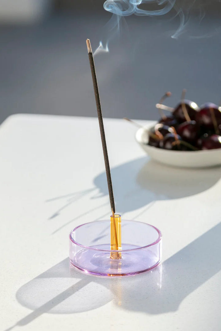 A Duo Tone Glass Incense Holder with a purple base and an orange holder, with an incense stick placed in it, situated on a white surface next to a white cup and some cherries.
