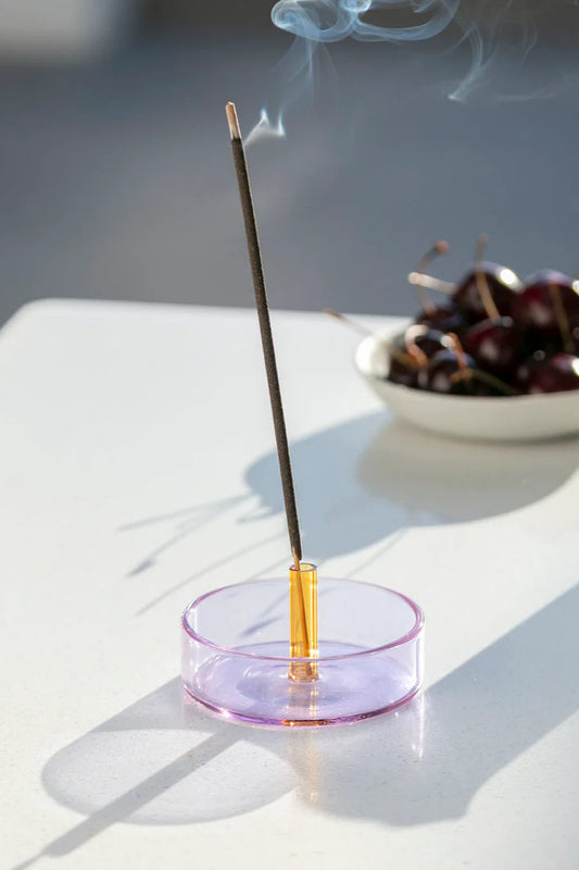 A Duo Tone Glass Incense Holder with a purple base and an orange holder, with an incense stick placed in it, situated on a white surface next to a white cup and some cherries.