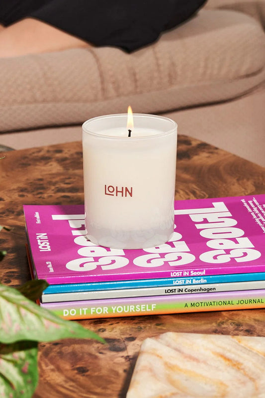 A white candle with the brand name 'LOHN' lit and placed on a table with a book and a plant in the background.