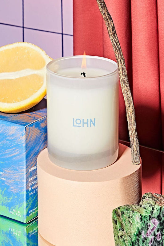 A candle with the brand name 'LOHN' lit on a circular beige platform, surrounded by citrus fruits and wooden blocks, on a background with a blue and red tiled wall.