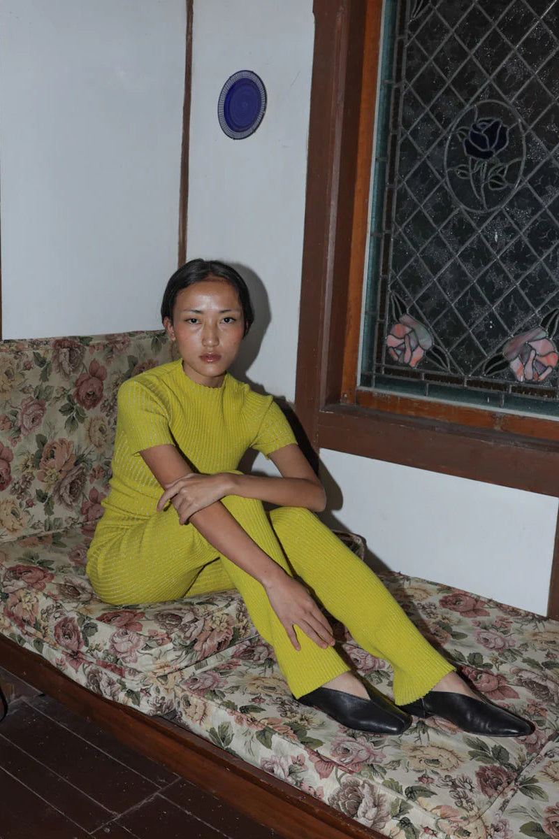 Person wearing a yellow outfit and black leather slipper shoes with a central seam and tulip shaped upper, a squared-off toe and 3/4" heel sitting on a floral-patterned couch.