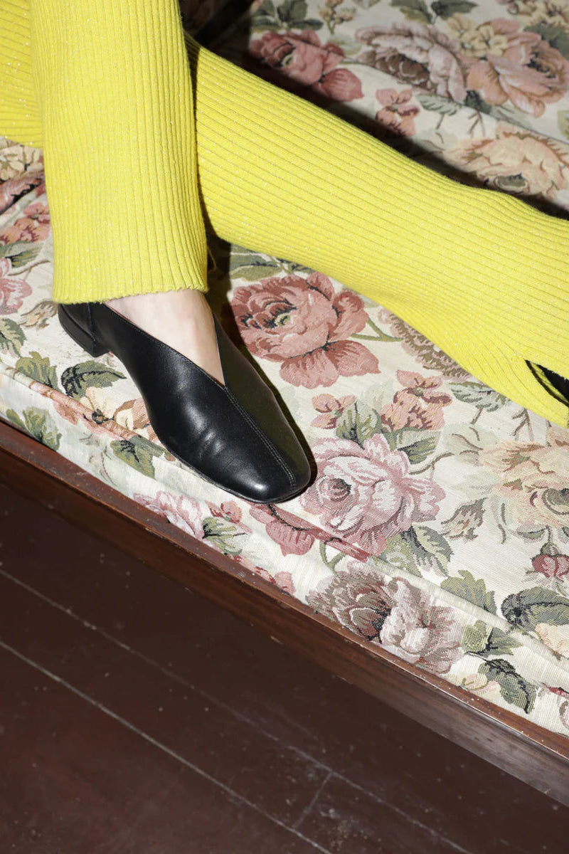 Person wearing yellow pants and black leather slipper shoes with a central seam and tulip shaped upper. Features a squared-off toe and 3/4" heel.