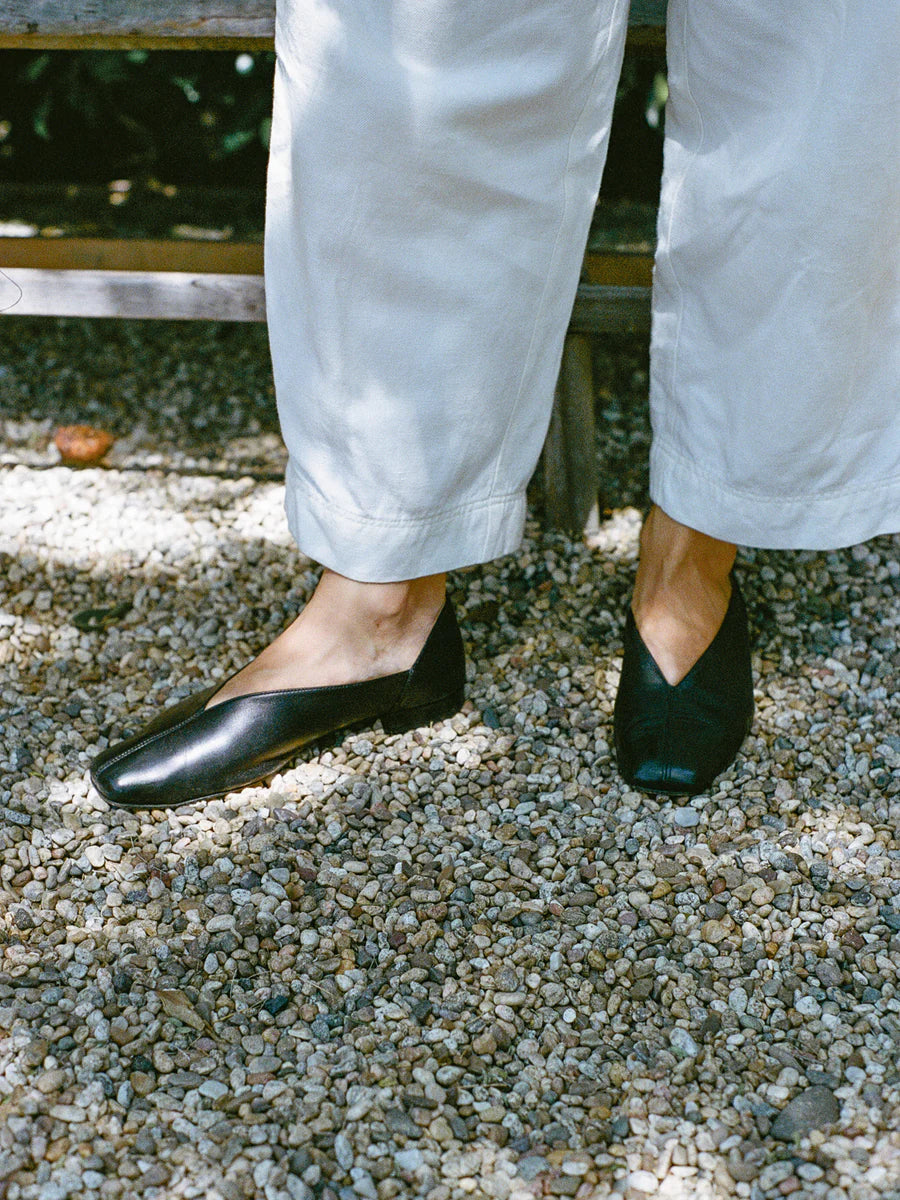 Person wearing black leather slipper shoes with a central seam and tulip shaped upper. Features a squared-off toe and 3/4" heel and white pants standing on a gravel surface.