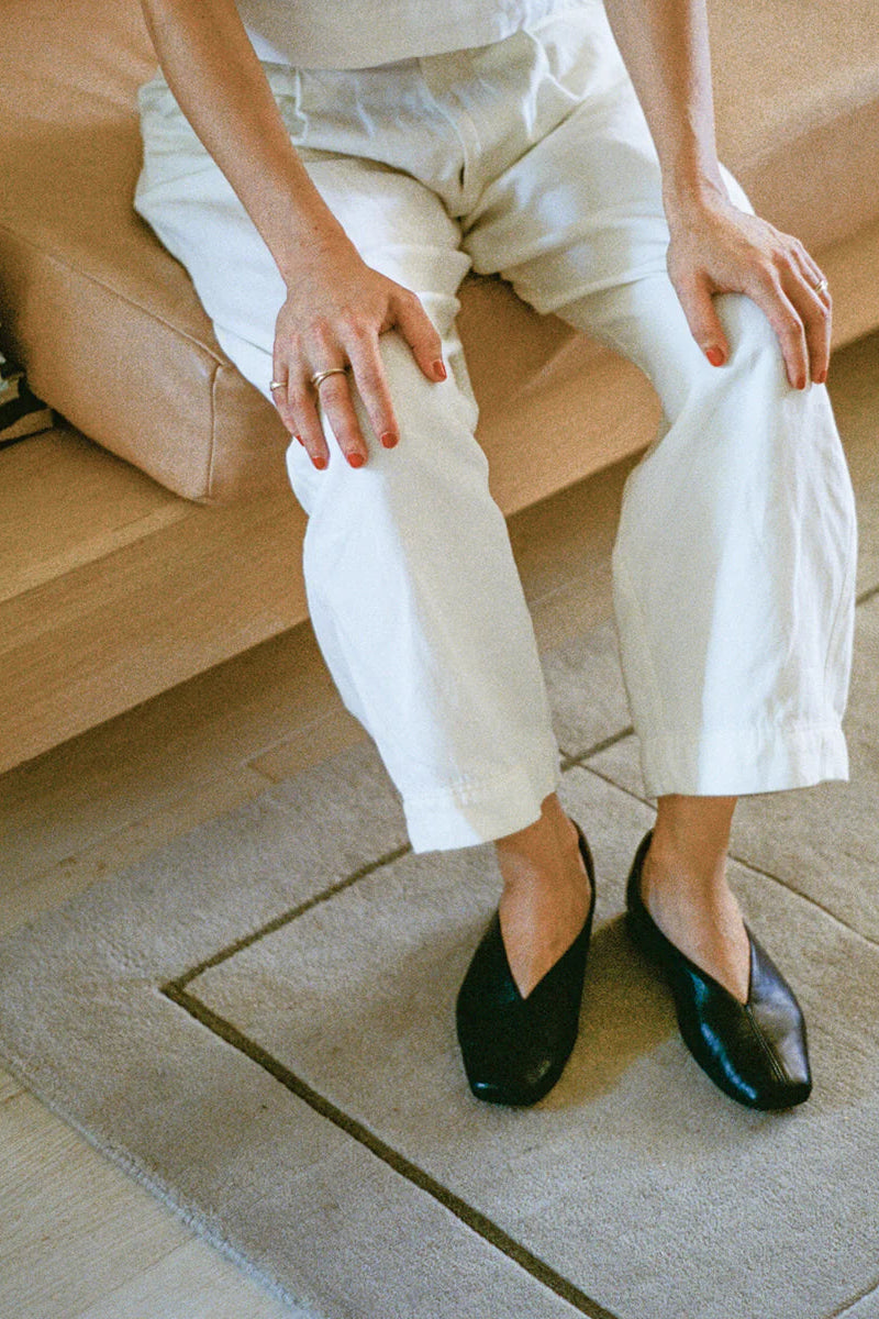 Person wearing white pants and black leather slipper shoes with a central seam and tulip shaped upper, a squared-off toe and 3/4" heel sitting on a couch.