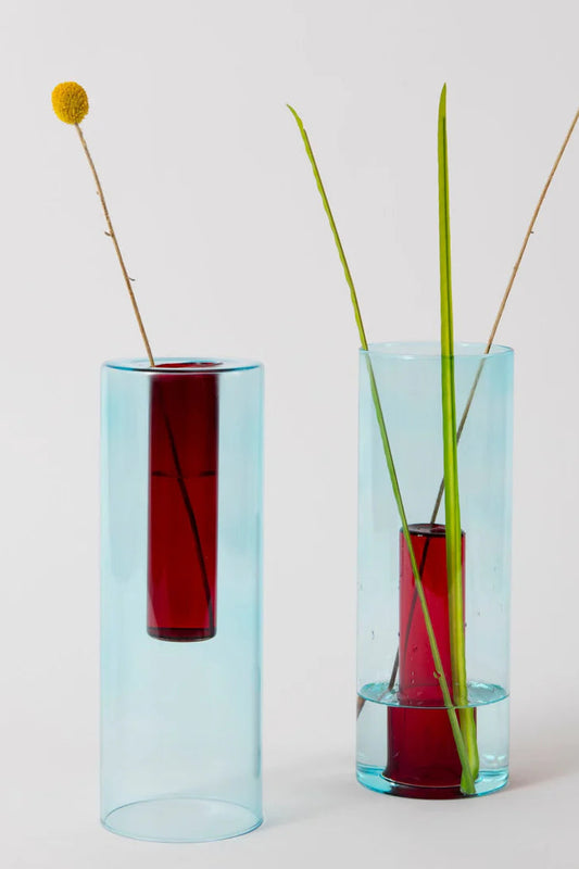 Two reversible glass vases, one with a red lower section and transparent upper, and the other with a transparent lower section and red upper. Each vase has a single stem of green plant matter in it, interacting visually with the colored sections of the vases.