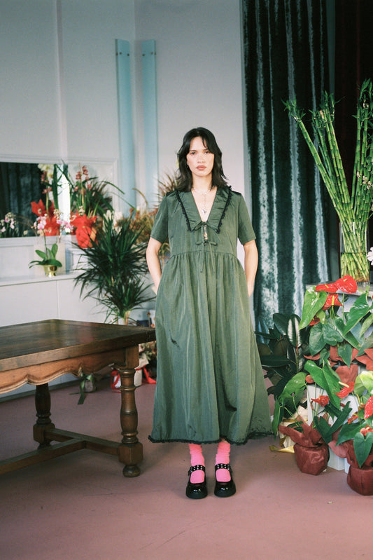 Woman in a green dress standing in a room with plants and a table.