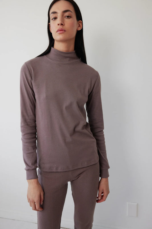 A woman standing against a white background, wearing a mushroom-colored turtleneck and leggings. The turtleneck has a ribbed neck and cuffs, and the leggings are also ribbed at the ankle.