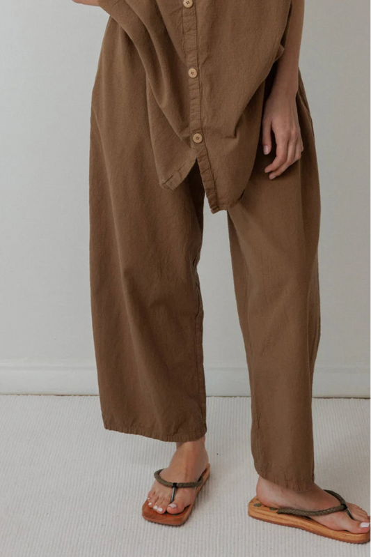 Person wearing a brown outfit with wide-leg pants and a buttoned shirt on a plain background