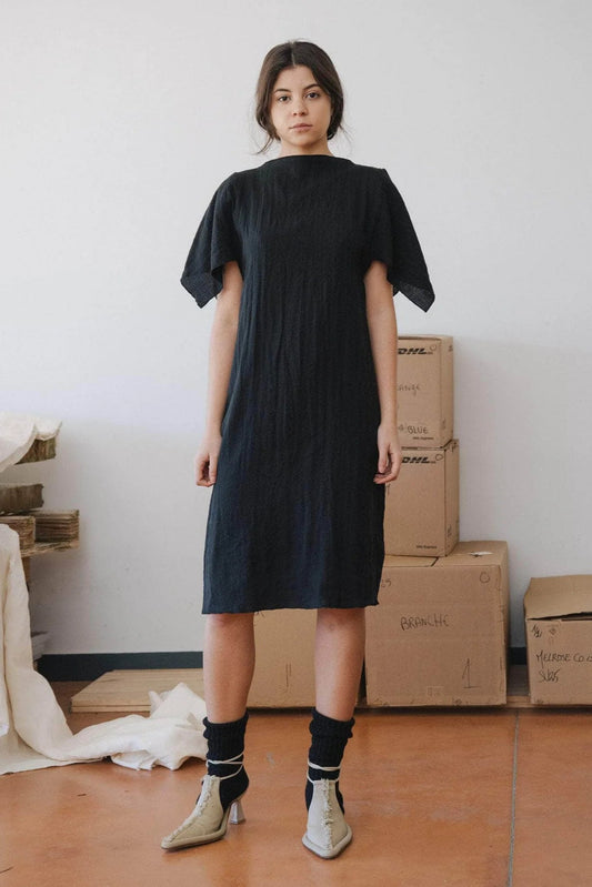 A model wearing a black crinkle linen dress with a boat neckline and slits on the sleeves at the shoulders. The dress reaches knee length and the model is standing in a room with cardboard boxes in the background.
