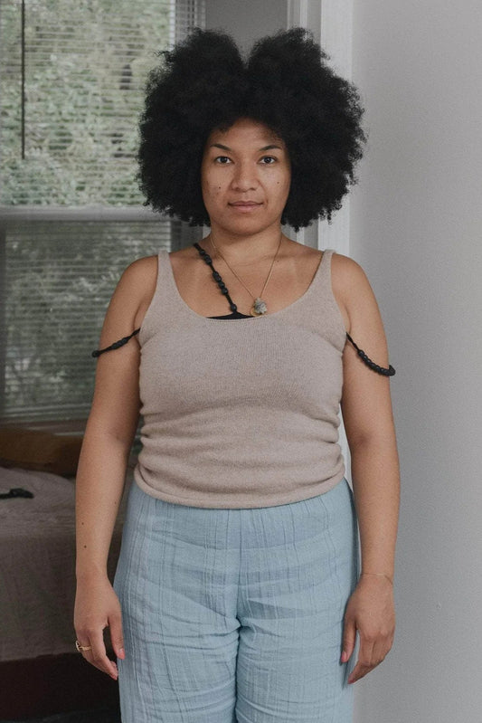 A person wearing a beige tank top with a scoop neck design, made of recycled cashmere and wool, standing in a room with natural lighting.