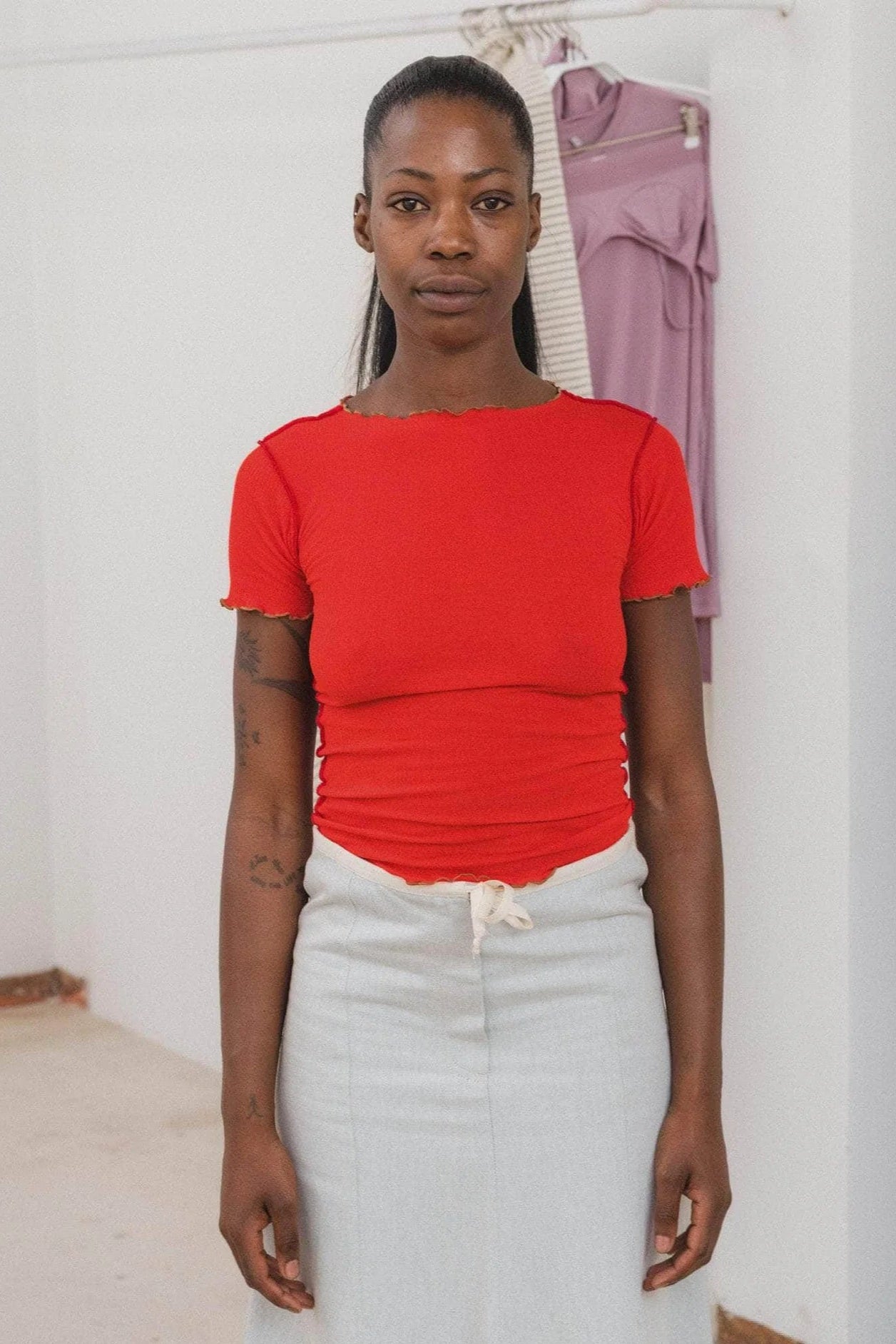 A person wearing a red boatneck tee shirt with contrast seams and lettuce edge at neckline and hems, standing in a room with a purple garment in the background.
