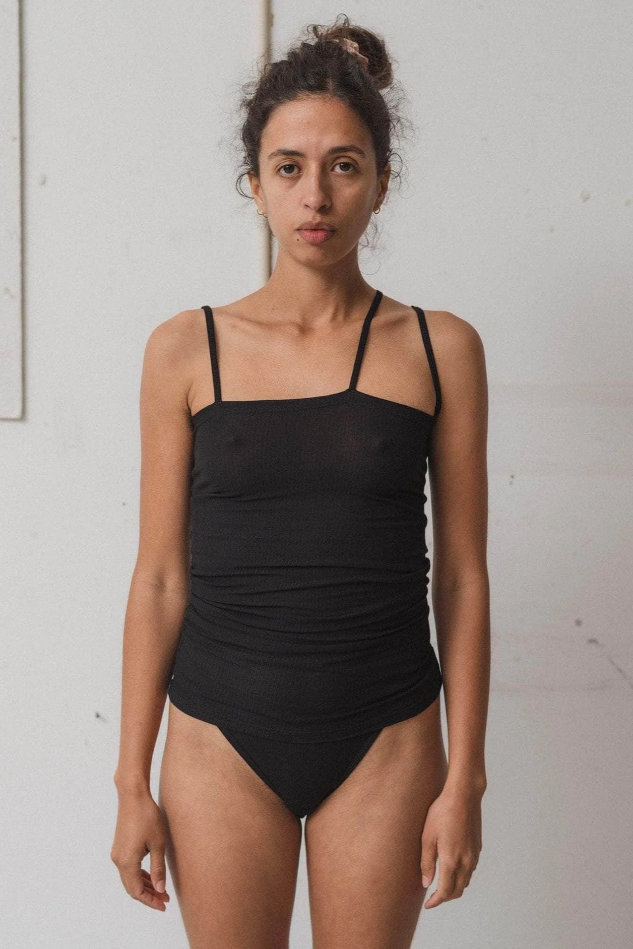 A model wearing a black tank top with thin shoulder straps and an asymmetrical strap, standing against a white background.