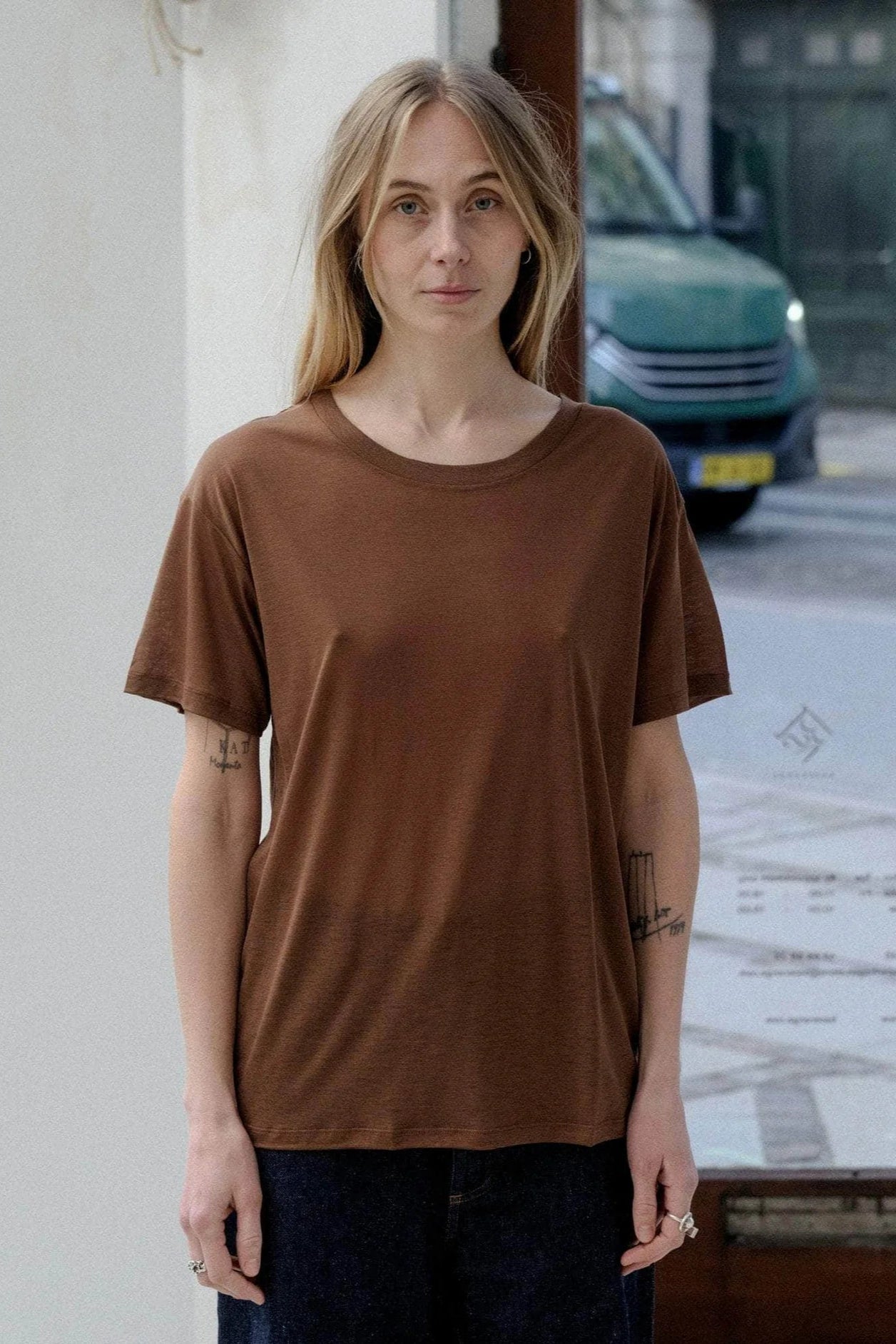 Person wearing a brown t-shirt standing on a street with a building and vehicle in the background.