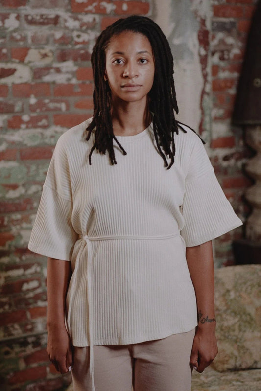 A person standing in a textured room wearing an off-white, ribbed cotton jersey short sleeve wrap top with a crewneck and dropped shoulder design.