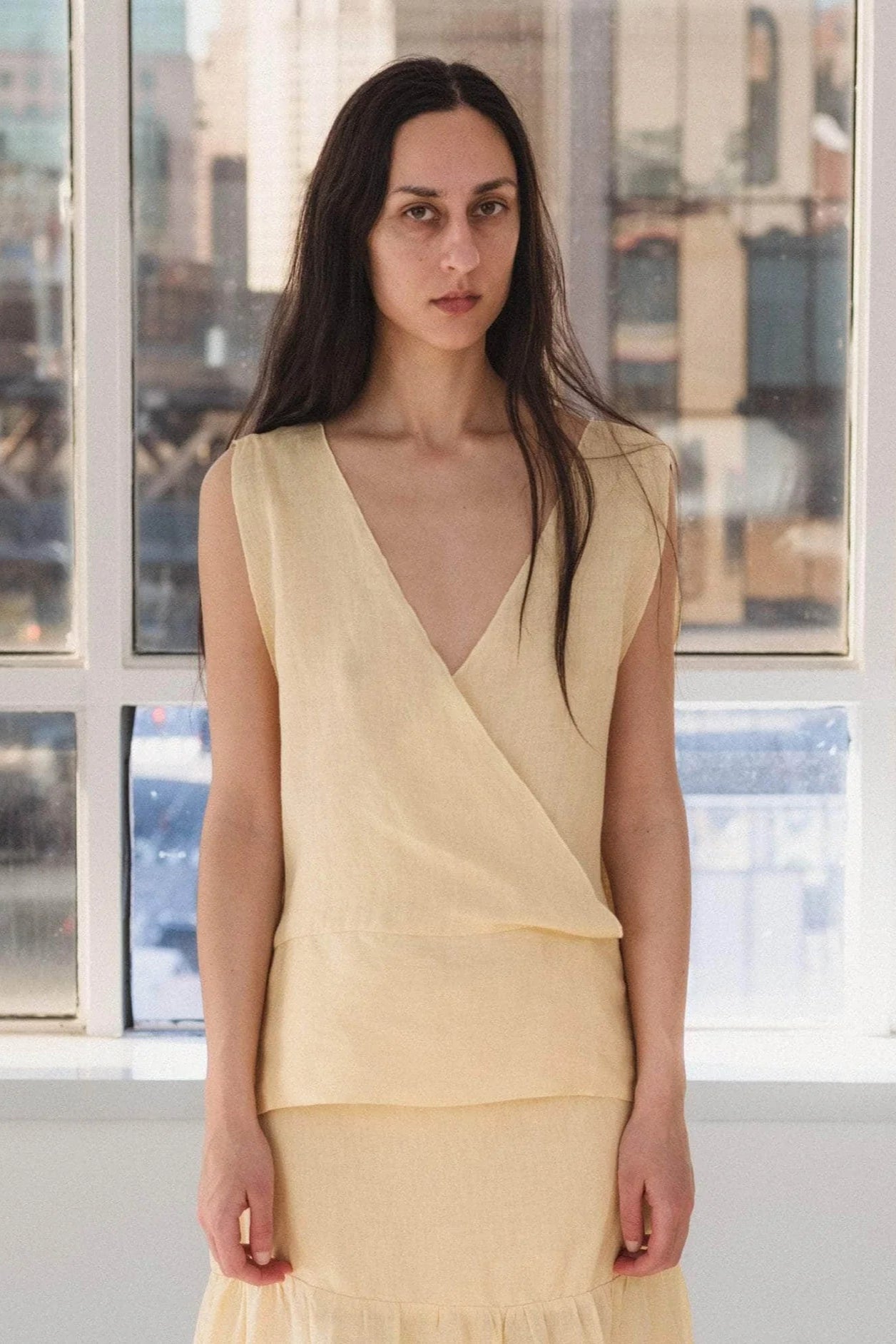 Woman wearing a light yellow top standing in front of large windows with a cityscape view.