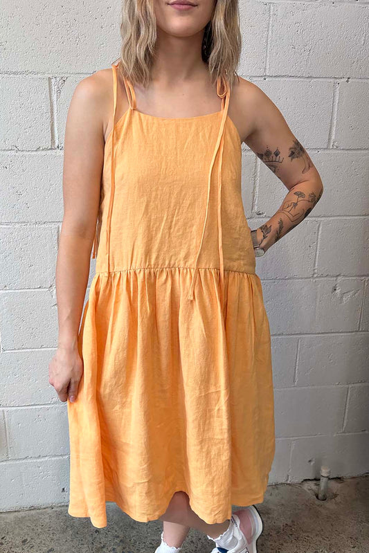 A person wearing a melon orange A-line linen dress with spaghetti straps and a full gathered skirt, standing against a brick wall.