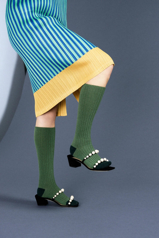 A person wearing green cashmere knee-high socks with a striped pattern and black shoes, standing against a grey background.