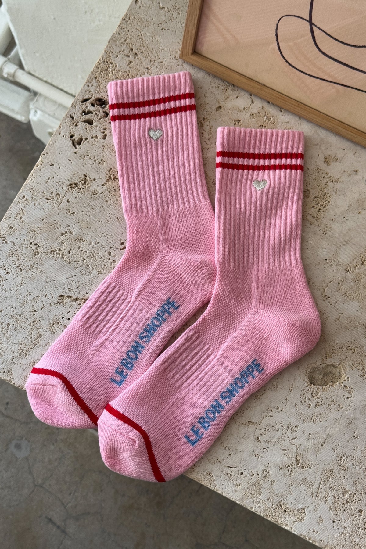 Pair of Le Bon Shoppe Embroidered Boyfriend Socks in amour pink laid flat on a travertine surface showing embroidered silver heart, red double stripe, sole branding and red contrast toe