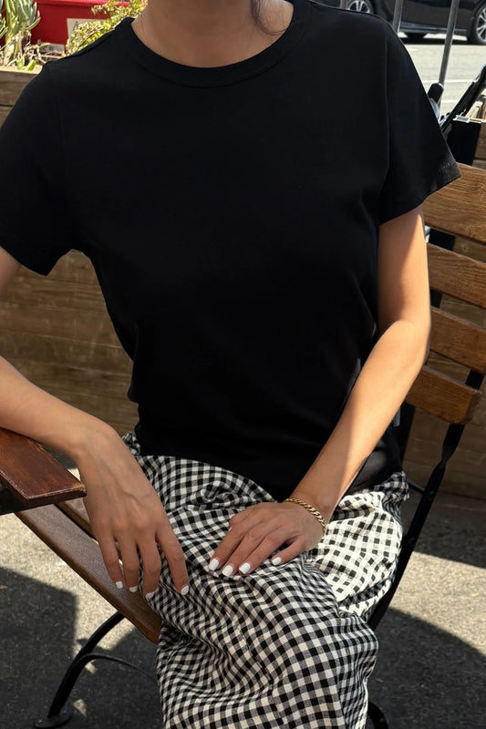 Le Bon Shoppe Go-To Tee in true black worn seated outdoors with black and white gingham trousers and a gold chain bracelet, showing relaxed fit in natural light