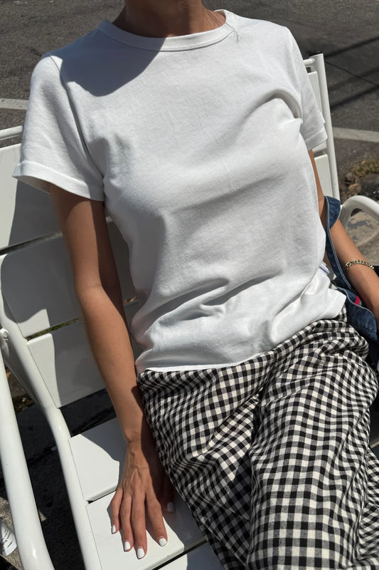 Le Bon Shoppe Go-To Tee in vintage white worn seated on a white chair in sunlight, styled with black and white gingham trousers and a denim bag