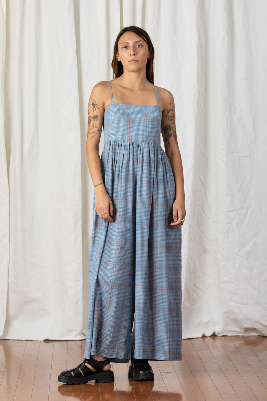 A person standing in front of a white curtain wearing a blue plaid full jumper with a square neckline and adjustable spaghetti straps.