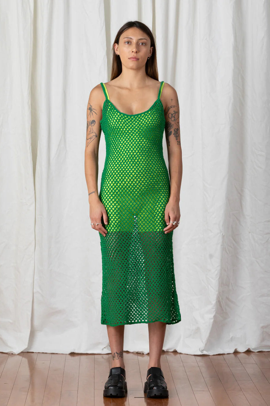A person wearing a green, open knit, cotton midi dress with spaghetti straps, standing in front of a white curtain.