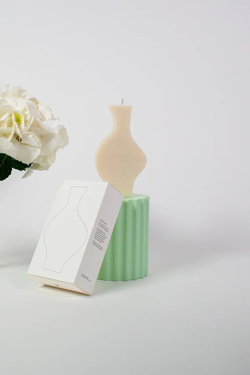 A white candle designed to resemble a vase, placed on a green surface with a box and white flowers in the background.