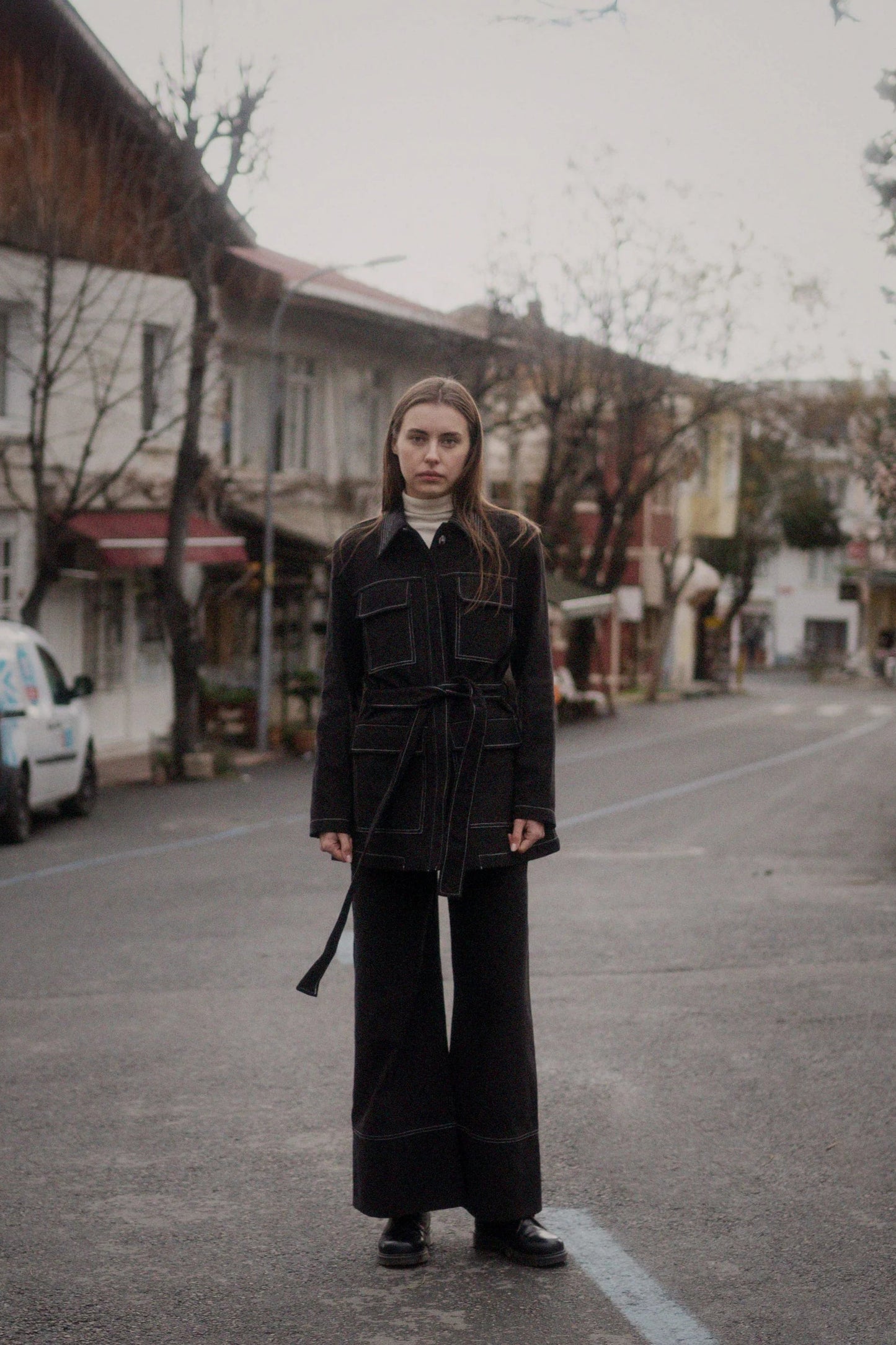 A person standing on a street wearing a black jacket with concealed button closure, breast and waist overlapping pockets, and a matching belt.