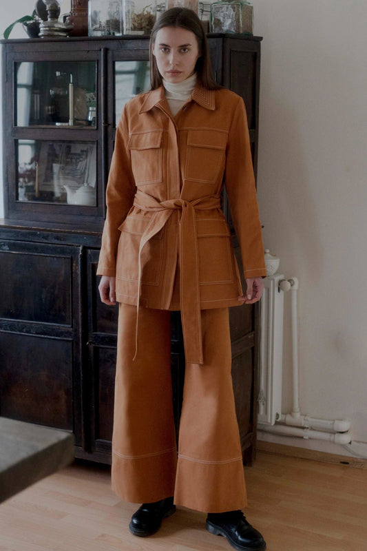 A person standing in front of a wooden cabinet wearing a brown jacket with concealed button closure, breast and waist overlapping pockets, and a matching belt.