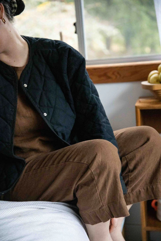 A person seated, wearing the Eva Pant in chestnut color, which is a wide-leg, high-waisted pant with deep pockets.