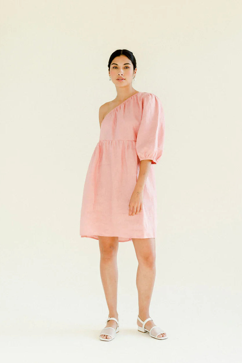 A woman standing in a pink one-shoulder loose babydoll dress with wide sleeve and elasticated hem, with the dress just below the elbow. She has an empire waist and side pockets, standing against a neutral background.