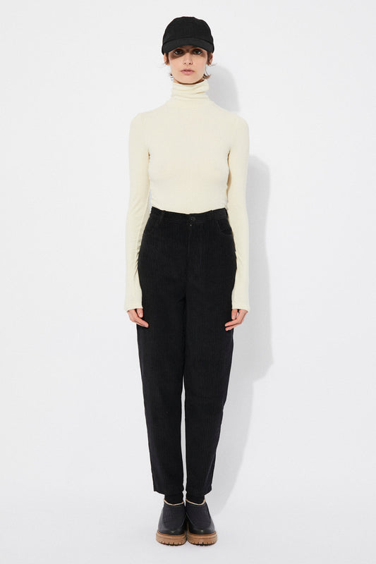 A model wearing high-waisted black corduroy pants with a balloon effect, featuring four pockets, a zipper closure, and button at the waist.