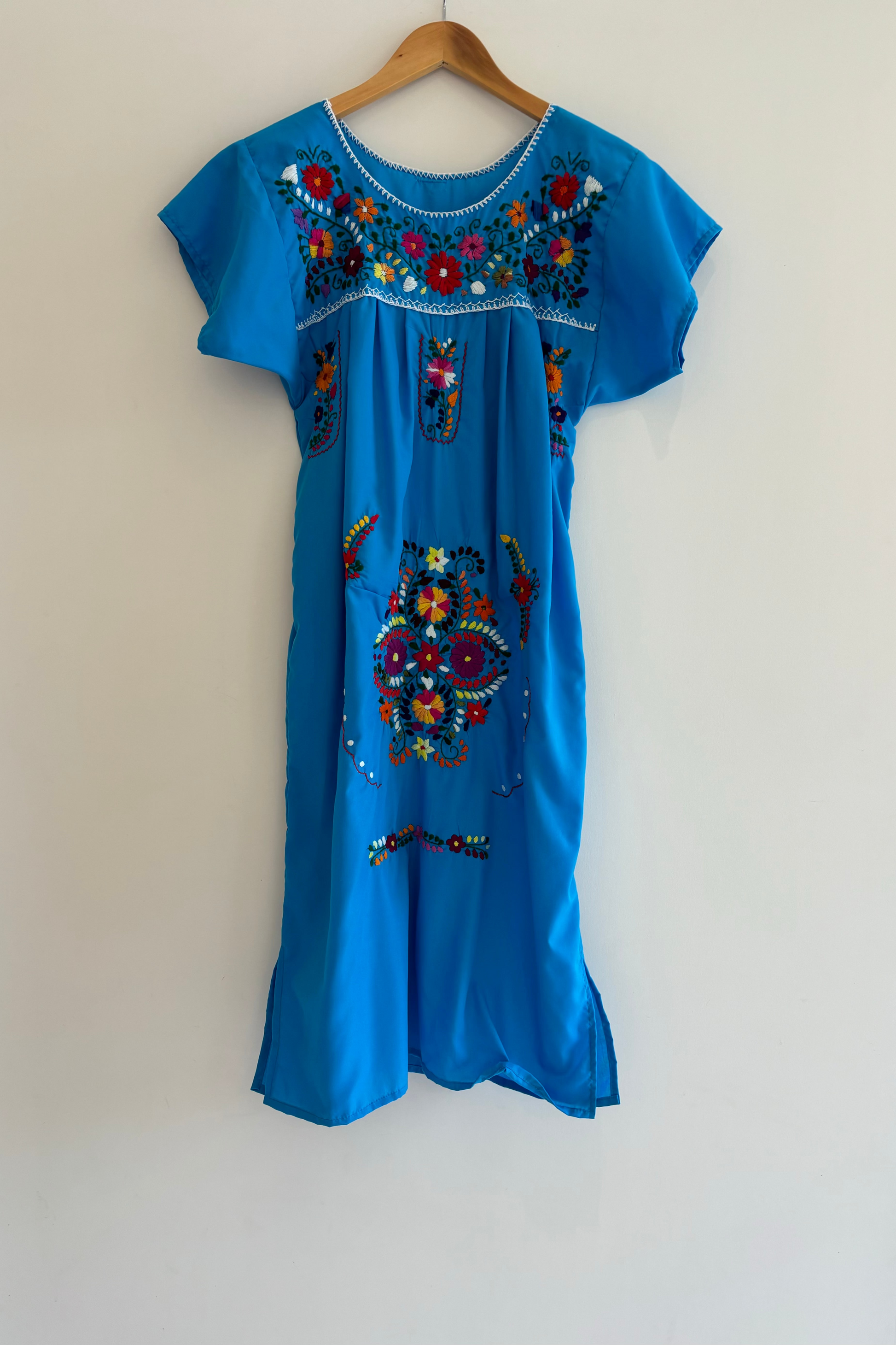 Blue dress with colorful embroidery on a hanger against a beige wall
