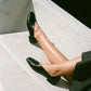 Person wearing black leather slipper shoes with a central seam and tulip shaped upper, squared toe and 3/4" heel on a light surface