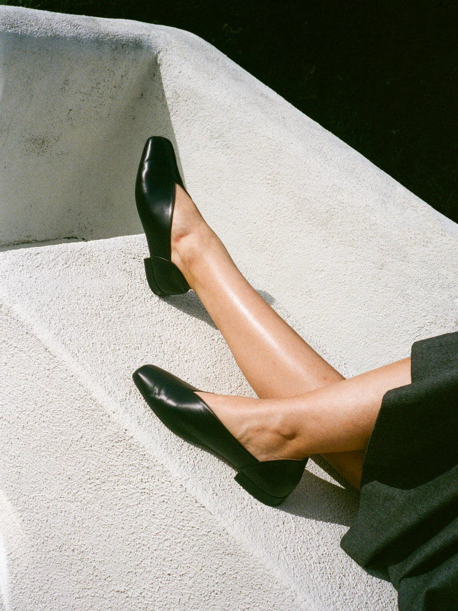 Person wearing black leather slipper shoes with a central seam and tulip shaped upper, squared toe and 3/4" heel on a light surface