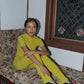 Person wearing a yellow outfit and black leather slipper shoes with a central seam and tulip shaped upper, a squared-off toe and 3/4" heel sitting on a floral-patterned couch.