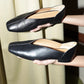 Black leather slipper shoes with a central seam and tulip shaped upper, squared-off toe and 3/4" heel held by a person against a neutral background
