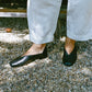 Person wearing black leather slipper shoes with a central seam and tulip shaped upper. Features a squared-off toe and 3/4" heel and white pants standing on a gravel surface.