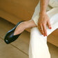 Person wearing black leather slipper shoes with a central seam and tulip shaped upper a squared-off toe and 3/4" heel wearing white pants sitting on a couch.