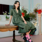 Woman in a green dress sitting on a wooden bench with plants in the background